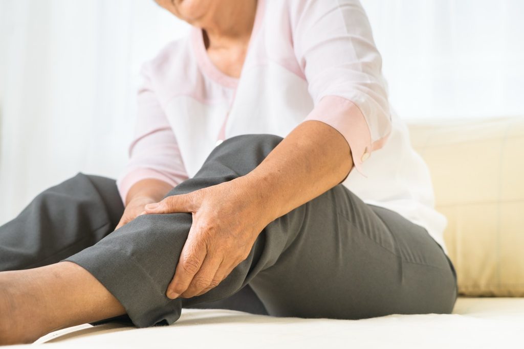 Senior woman holding her leg in pain while sitting at home, illustrating potential circulation issues or leg discomfort.